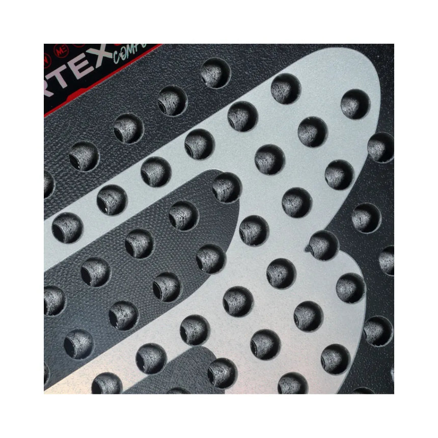 Close-up of a textured surface with raised black circles on a silver background, reminiscent of the Bullpadel Vertex 04 Comfort 2024 Martín Di Nenno design. A fragment of a black and red logo is visible in the top left corner.