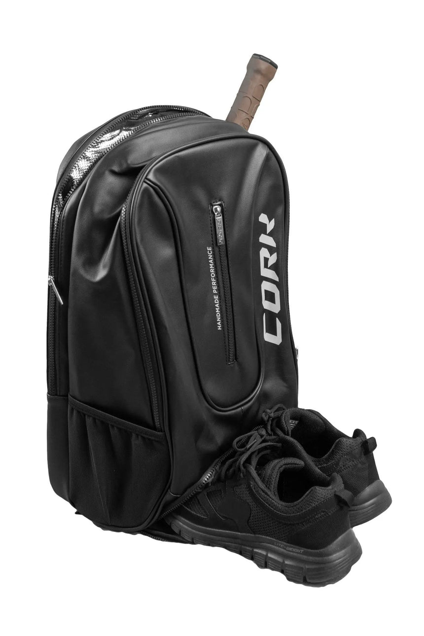 A Cork Padel backpack in black, tailored with multiple compartments, zippers, and a thermal pocket for your essentials, with a pair of black athletic shoes and a tennis racket handle peeking out—ideal as a lightweight padel backpack.