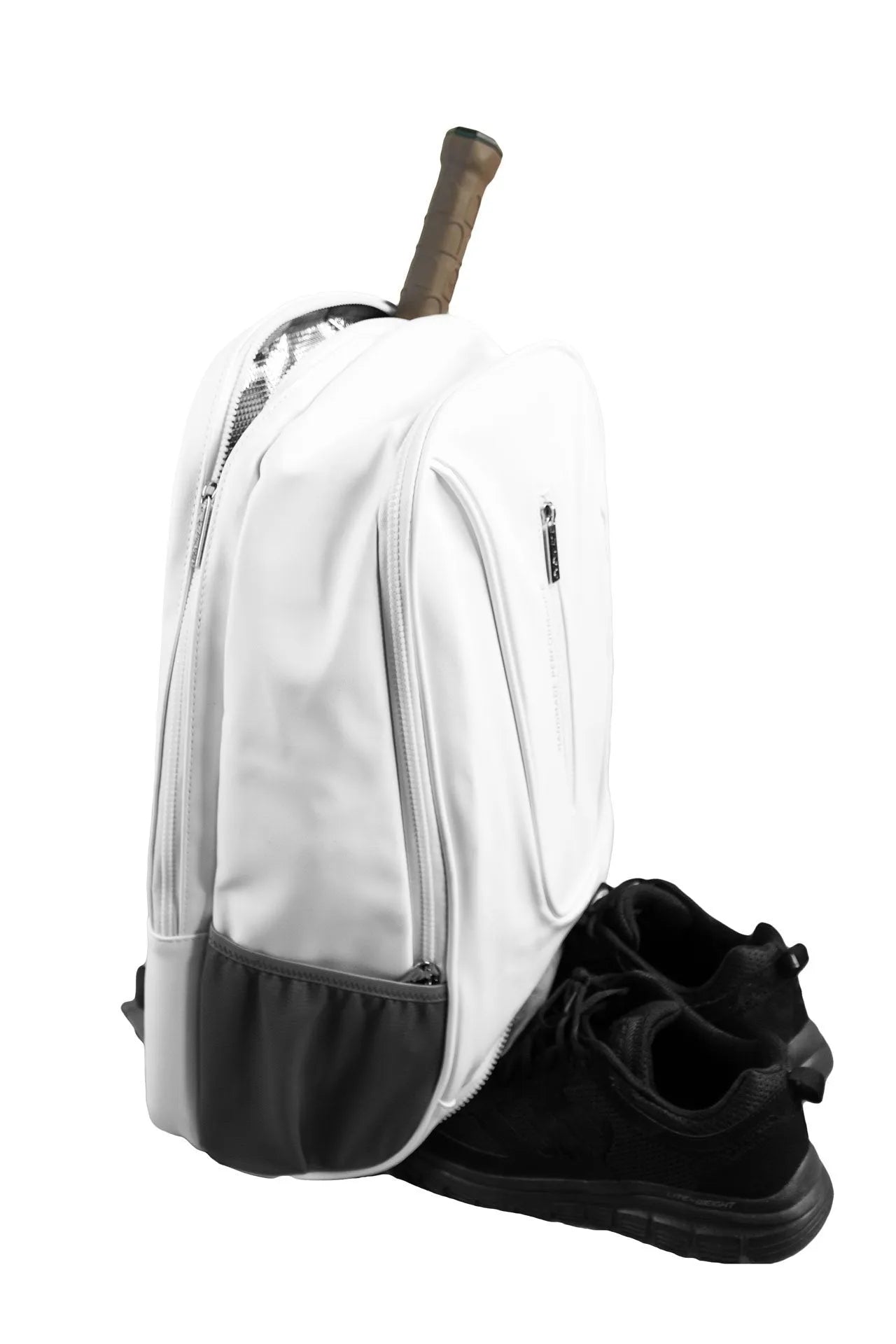 The Cork Padel Backpack, by Cork Padel, in white with black trim, features a tennis racket handle sticking out and a thermal pocket for gear protection. A pair of black athletic shoes is placed slightly in front of the lightweight backpack.