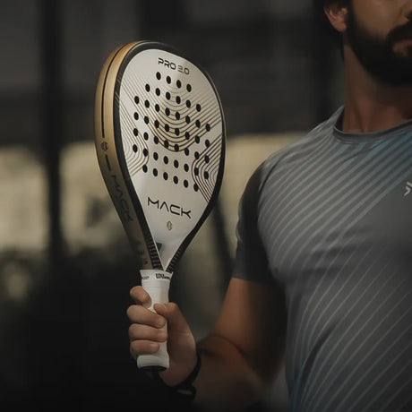 A person is holding a teardrop-shaped Mack Pro 2.0 2024 White racket by Mack Starke, featuring a unique pattern and aluminized Textreme, while wearing a gray sports shirt against a blurred background.