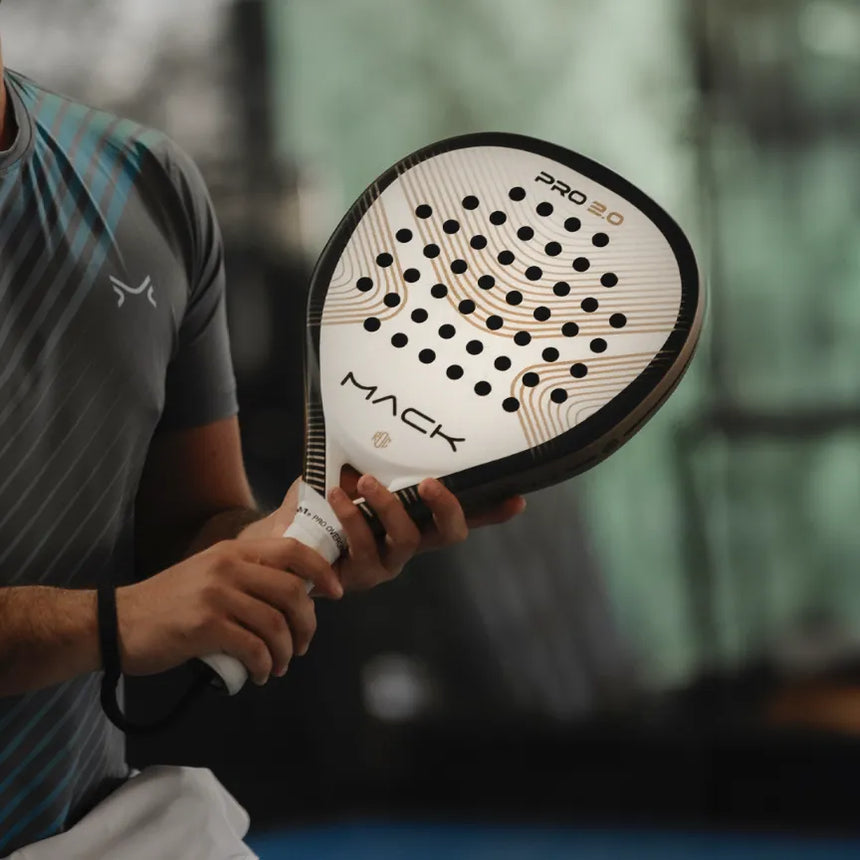 An individual is holding a Mack Starke Mack Pro 2.0 2024 White padel racket featuring a teardrop shape and an aluminized Textreme finish with a white grip, wearing a patterned gray shirt in an indoor sports facility with a blurred background.