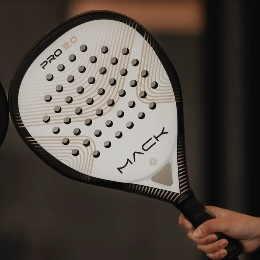 A person holds a teardrop-shaped padel racket with MACK boldly emblazoned on it. This Mack Starke Mack Pro 2.0 2024 White, aluminized and featuring patterned holes and curved lines, stands out against the blurred background.