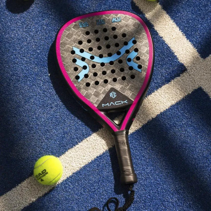 The Mack Starke I-Pro Titanium 2024 racket, featuring a pink and grey design with advanced Black Soft EVA rubber, lies on a blue court with white lines. A nearby yellow tennis ball rests as shadows faintly cast over the surface.