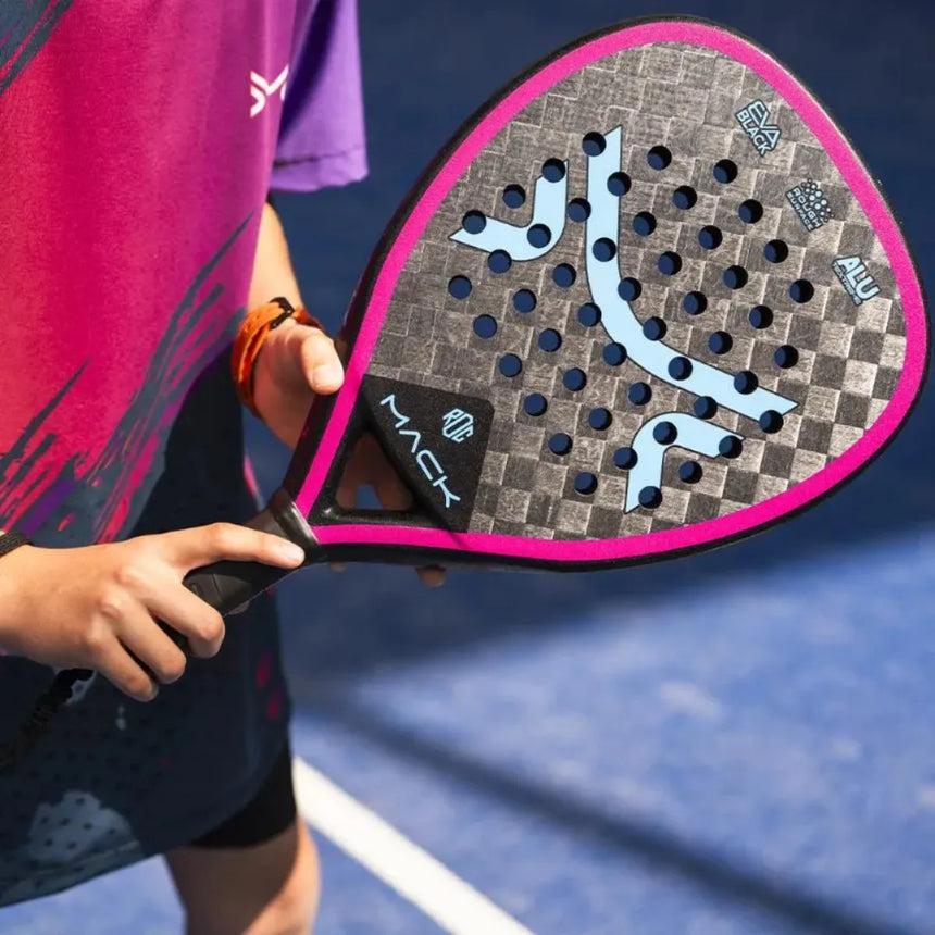 A person holds the Mack Starke I-Pro Titanium 2024 padel racket, highlighting its diamond shape and Black Soft EVA rubber on a vibrant blue court. This pink-and-black racket, featuring brand logos, contrasts perfectly with their purple sports shirt.
