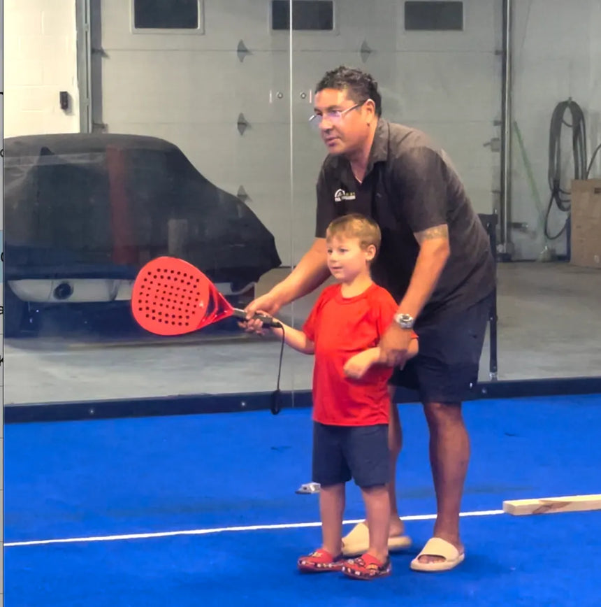 On a blue indoor sports court, a man assists a young boy holding a red paddle during a Padel Smash clinic. They focus on the swing with covered objects and other sports gear visible in the background.