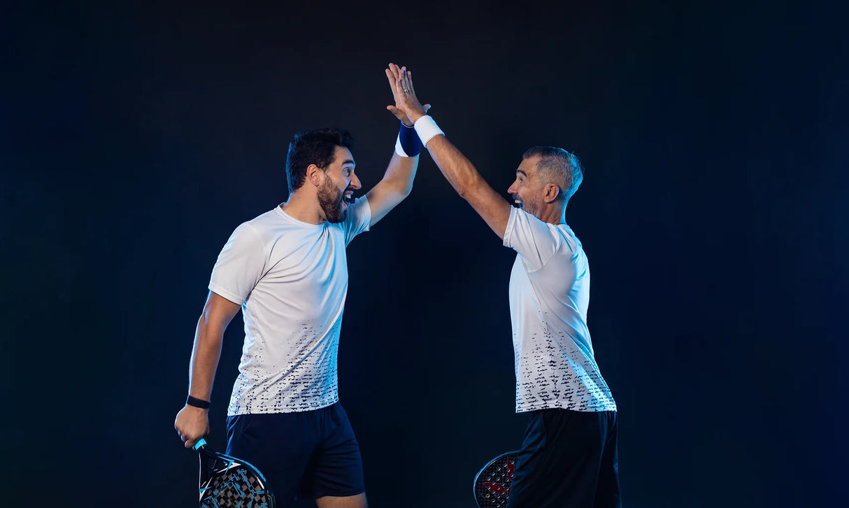 Two men in white shirts and dark shorts high-five against a dark background, each holding a paddle, symbolizing an energetic and celebratory moment, potentially related to Padel Consulting by Padel Smash Academy.