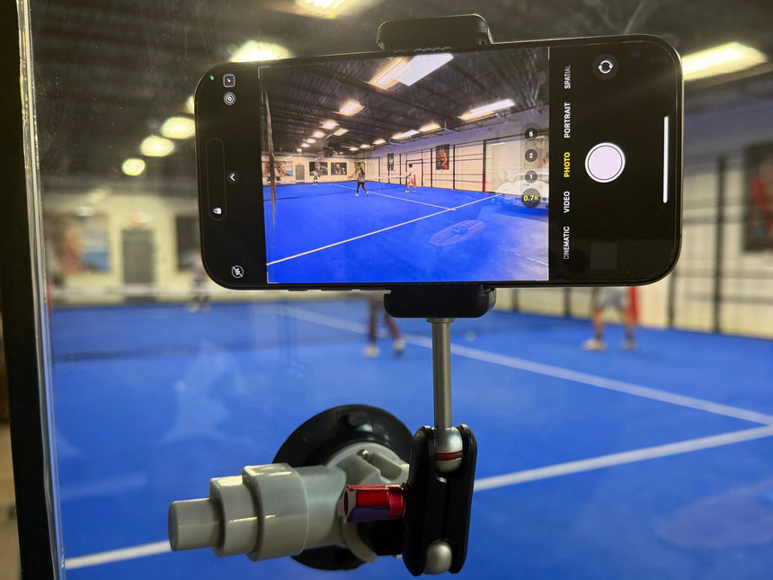 A smartphone on the Padel Smash Academys Padel Pro Mount attached to a glass pane captures the excitement of a pickleball game, highlighting dynamic movement as it tracks two players on a blue court inside a gym.
