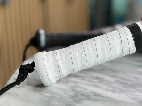 The close-up features a Padel Racket Silicone Ring by Padel Smash Academy, wrapped in white tape for improved grip and wear prevention, with a black wrist strap attached. The blurred background highlights a light surface and parts of another racket.