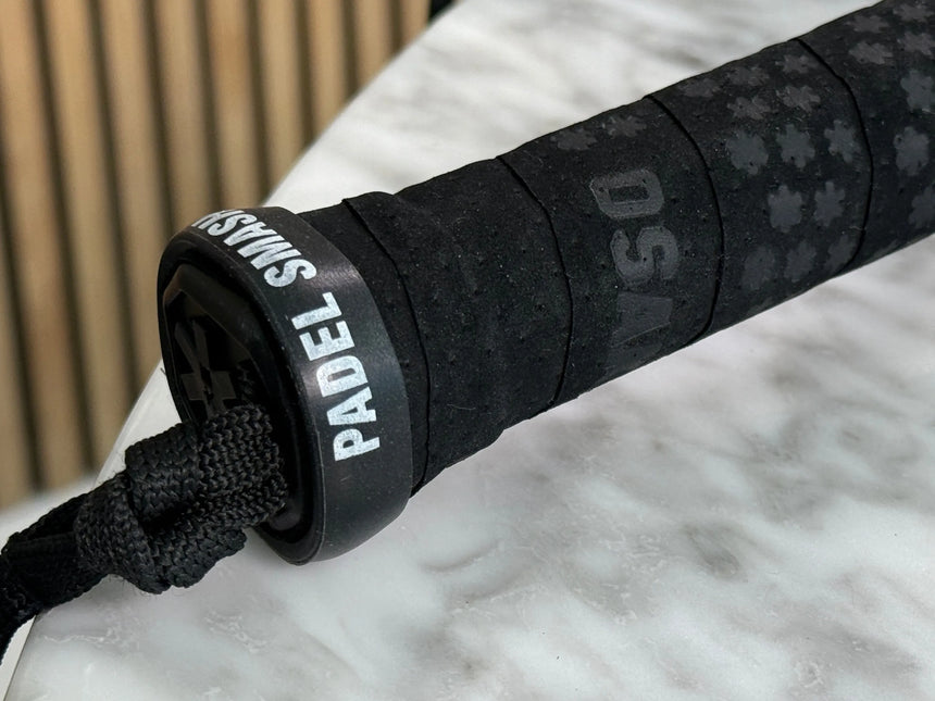 Close-up of a black padel racket handle featuring embossed designs and a Padel Racket Silicone Ring x2 by Padel Smash Academy to prevent grip wear. The strap reads Padel Smash. It rests on a light gray marble table with a blurred wooden panel backdrop.
