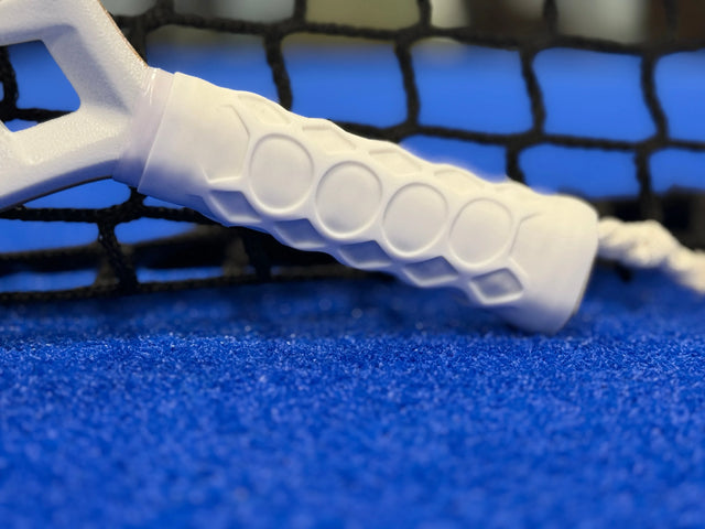 A white pickleball paddle handle with the Power Grip by Padel Smash Academy for added comfort leans against a black net on the blue court surface.