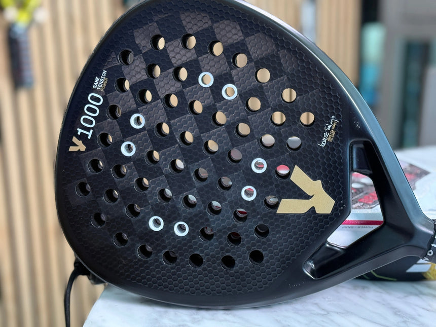 A close-up of the black Racket Anti-Vibration System by Padel Smash Academy showcases a checkered pattern with circular holes, gold accents, and the text 1000. Its displayed on marble against blurred wooden paneling, ideal for padel athletes.