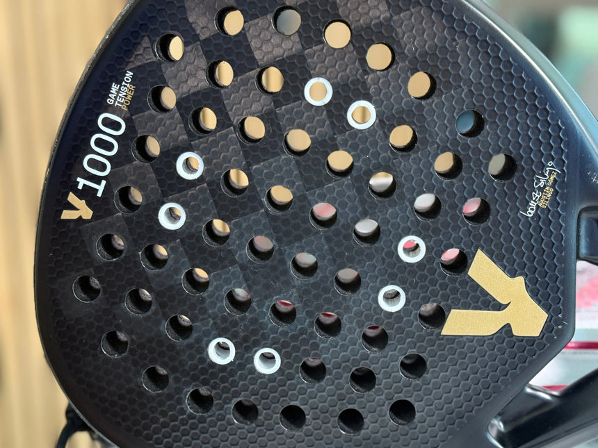 Close-up of a Padel Smash Academy Racket Anti-Vibration System with a black, perforated surface and golden arrow design. 1000, single, and composite are visible. Ideal for athletes seeking enhanced performance. Background is blurred.