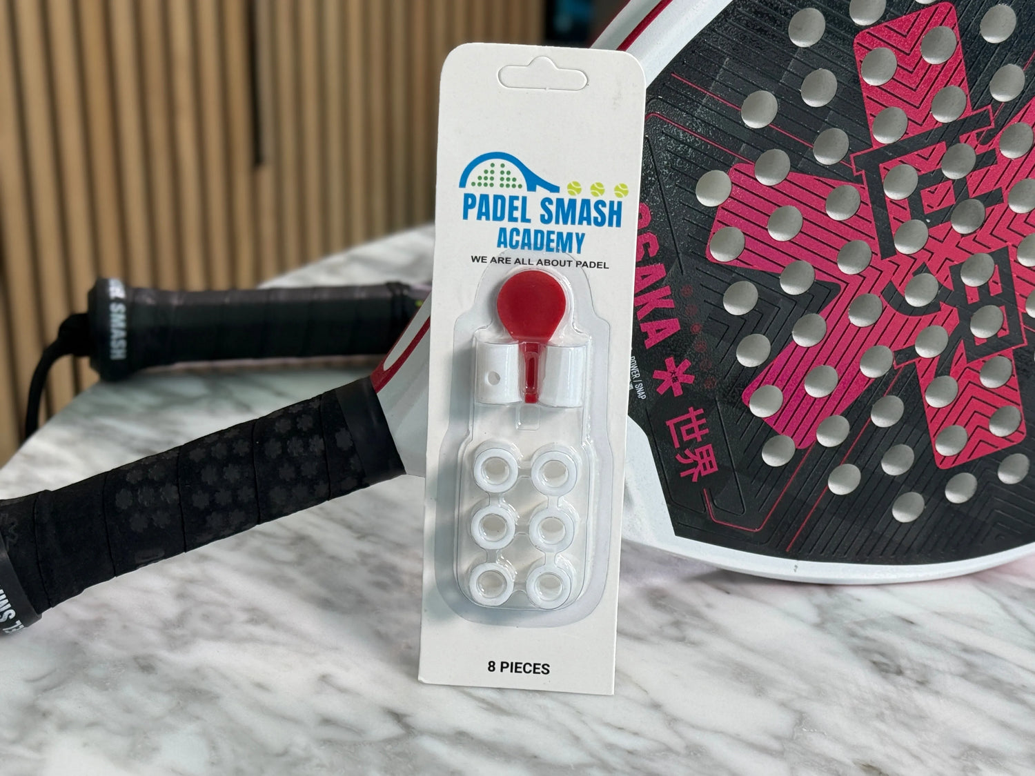 A padel racket sits on a marble surface alongside a Padel Smash Academy Racket Anti-Vibration System, tailored for athletes. The package contains six clear anti-vibrators and one red cap.