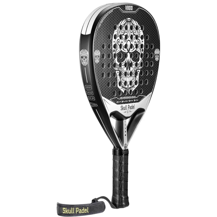 The SKULL PADEL V800 - 1K WHITE features a black and silver design with an intricate skull on the carbon surface. It has a black grip with Skull Padel wrist strap and EVA rubber for comfort, while the model name V800 is marked at the top. Brand: Skull Padel.