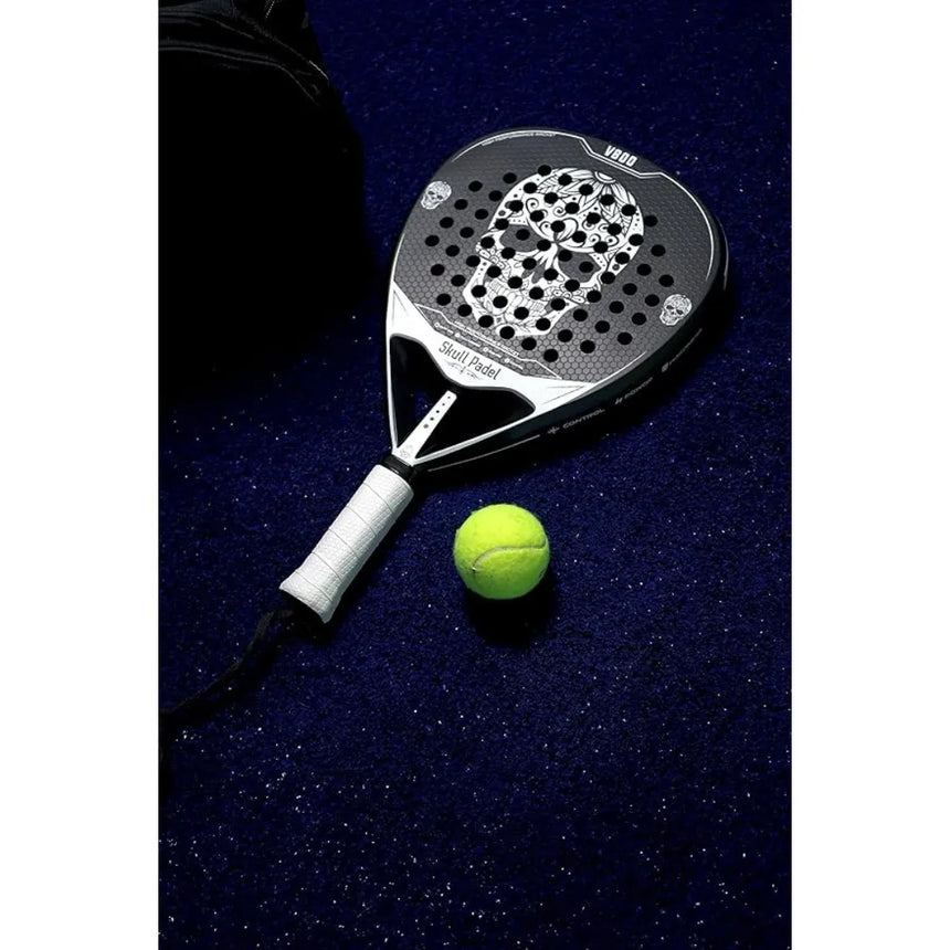 The SKULL PADEL V800 - 1K WHITE (LIKE NEW) racket by Skull Padel, featuring a vibrant skull design and durable carbon surface, rests on the dark blue court with a yellow padel ball.