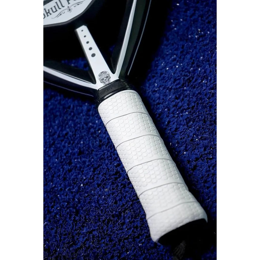 Close-up of a SKULL PADEL V800 - 1K WHITE (LIKE NEW) paddle tennis racket handle with a white grip on a textured blue surface. The black frame, made from premium carbon, displays the partially visible Skull Padel logo above the handle.