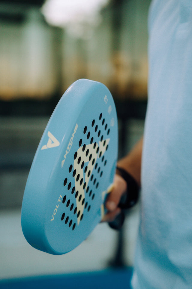 Close-up of someone holding an Antone Padel Volti racket featuring 12K carbon construction and black perforations, wearing a white shirt with a blurred indoor court background.