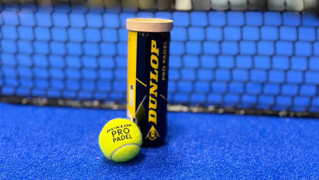 A Padel Smash tube of Balls Split by Player and a tennis ball sit on a blue court, with a padel net in the background. This shared brand enhances the padel theme, perfect for four players sharing the cost of balls.