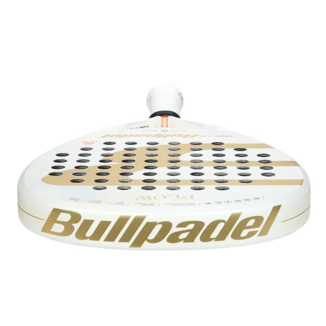 Close-up of the Bullpadel Racket Flow W 2024 Alejandra Salazar by BullPadel, showcasing its white color with gold branding. It features a Fibrix hybrid fiber surface with black circular holes and a handle equipped with the Vibradrive system plus an orange accent for better grip.