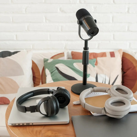 A home podcast setup for Padel enthusiasts features a black microphone from Padel Smash Academy, two headphones, a closed laptop, and a notebook with a pen on a round wooden table. A couch with decorative pillows sits in the background against a white brick wall.