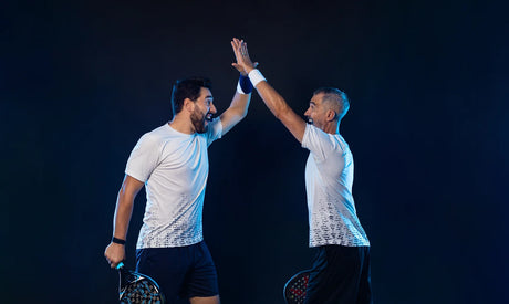 Two men in white shirts and dark shorts high-five against a dark background, each holding a paddle, symbolizing an energetic and celebratory moment, potentially related to Padel Consulting by Padel Smash Academy.