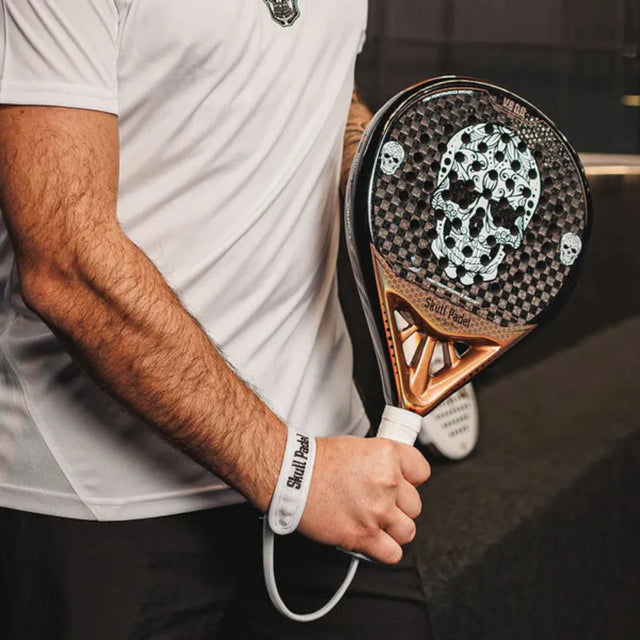 A person wearing a white shirt and wristband holds the SKULL PADEL V600 - 12K COPPER SKULL racket by Skull Padel, featuring a black and white skull design. The grip is black and orange, ideal for intermediate players. The dark background enhances this game-preparation scene.
